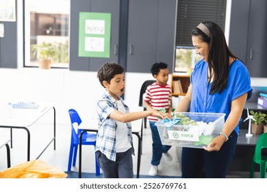 In school, in classroom, biracial female teacher and two young boys recycling. She wearing blue top, boys in colorful t-shirts, all focused on a bin, learning, education, unaltered - Powered by Shutterstock