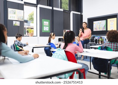 In school, in a classroom, a young biracial female teacher is explaining a lesson. Diverse students are listening, some raising hands to answer, unaltered. - Powered by Shutterstock