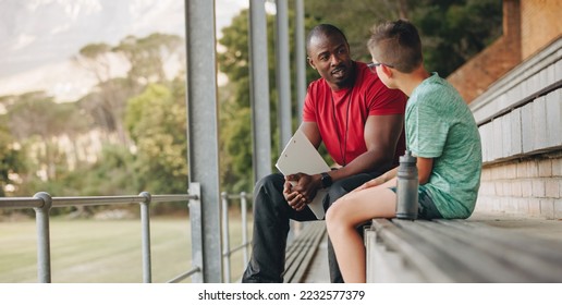 School coach talking a young student while sitting outside. Physical education teacher motivating a young boy in elementary school. Child mentor giving support and guidance. - Powered by Shutterstock