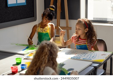 In school, during art class, diverse young students painting on canvas. Biracial girl with bun, wearing yellow, talking to friend with curly hair, unaltered - Powered by Shutterstock