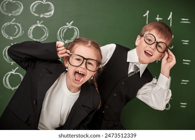 School education. Two smart schoolchildren - a boy and a girl - in a school uniform and glasses, cheerfully pose at the blackboard in the classroom. - Powered by Shutterstock