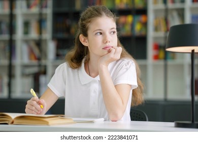 School girl wearing white t-shirt sitting at table and writing in exercise book, looking to side and daydreaming, bookshelves on blurred background. Pupil doing assignment in class