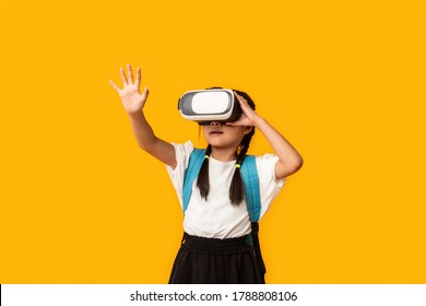 School Innovations. Asian Schoolgirl Wearing Virtual Reality Glasses Experiencing VR Standing Over Yellow Background. Studio Shot - Powered by Shutterstock