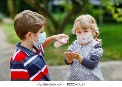 School Kid Boy Helping Little Toddler Sister Cleaning Hands With Sanitizer Spray. Brother And Cute Little Girl In Medical Mask Learn Hygiene Rules. Family During Coronavirus Pandemic Quarantine