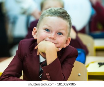 School Kid In Class. Funny Little Boy Pupil Smiling While Sitting In Desk In Suit. Inquisitive, Interested With Eyes Wide Open.