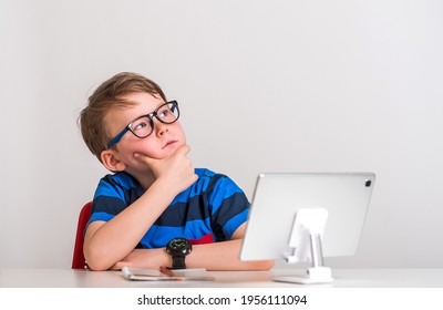 School Kid In Glasses Sitting At The Table Using Tablet Laptop Computer For Online Lesson. Thinking Upset Child With Hand On Head For Mistake, Remember Error. Forgot, Bad Memory Concept.