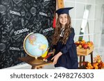 School Kid in graduate headdress is studying in classroom against of blackboard. Smiling little girl in a school uniform and master