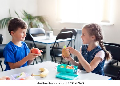 School Kids Eating Healthy Food Together. Happy Children Sitting At Table With Packed Lunch Boxes. Back To School Concept. Boy And Girl Treat Each Other Fruits.