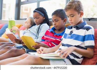 School Kids Sitting On Sofa And Reading Book In Library At School