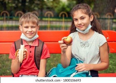 School learners in medical masks sitting on the bench in school yard having a snack during a break. Back to school concept