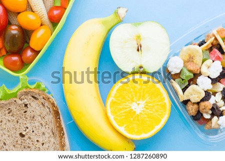 School lunch box. Bread, apple, candies, baby corns, carrot and tomatoes in green plastic container. Top view, blue background