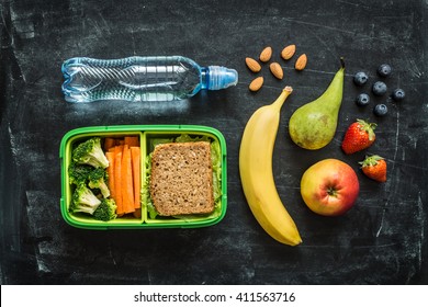 School Lunch Box With Sandwich, Vegetables, Water, Almonds And Fruits On Black Chalkboard Background. Healthy Eating Habits Concept. Flat Lay Composition (from Above, Top View).