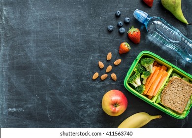 School lunch box with sandwich, vegetables, water, almonds and fruits on black chalkboard. Healthy eating habits concept - background layout with free text space. Flat lay composition (top view). - Shutterstock ID 411563740