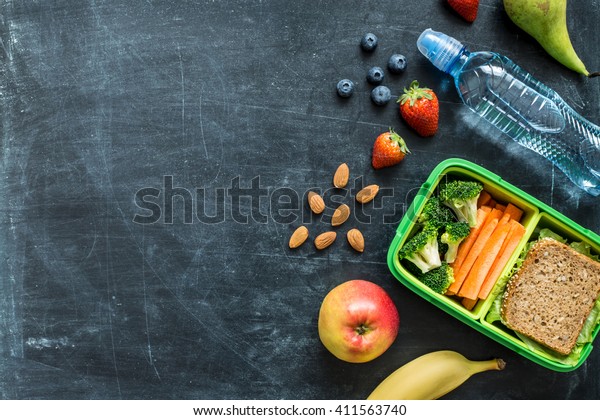 School lunch box with sandwich, vegetables, water,\
almonds and fruits on black chalkboard. Healthy eating habits\
concept - background layout with free text space. Flat lay\
composition (top view).