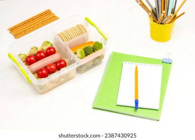 School lunchbox on table. Notebook pen, and bottle of yogurt. Yellow basket with pencils and edible straws. White background. Top view