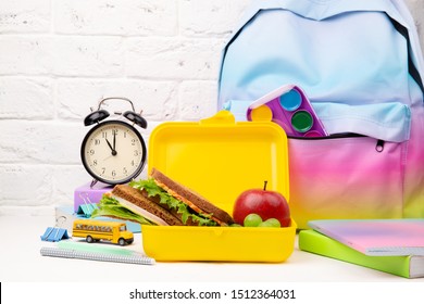 School Objects, Books, Alarm, Bag And Yellow Lunch Box With Sandwich, Apple And Carrot On Brown Wood Table. Copy Space.