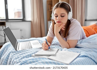 school, online education and e-learning concept - bored teenage student girl in headphones with laptop computer writing to notebook lying on bed at home