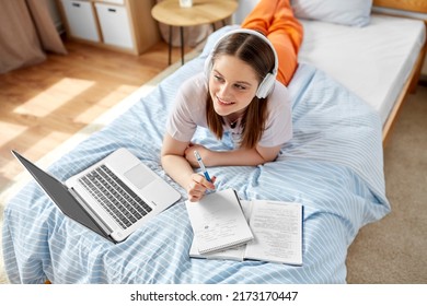 Schule, Online-Bildung und e-Learning-Konzept - glückliches, lächelndes Schülermädchen in Kopfhörern mit Laptop-Computer-Schreibmaschine zum Notebook im Bett zu Hause