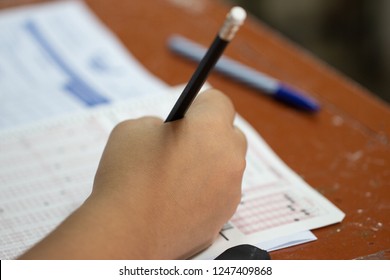 School Students Hands Taking Exams, Writing Examination With Holding Pencil On Optical Form Of Standardized Test With Answers Sheet On Desk Doing Final Exam In Classroom. Education Assessment Concept