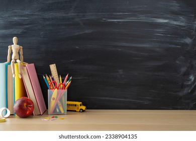 School supplies layout. Side view photo of desk setup with pencils organizer, ruler, books, red apple, mannequin body, and more on chalkboard background. Great for educational content or advertising