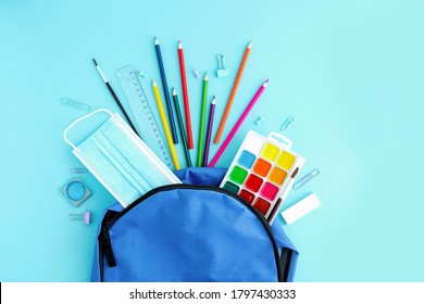 School supplies and a protective medical mask poured out of a blue backpack or knapsack on a light background, top view. Back to school, study after vacation and quarantine. Place for text