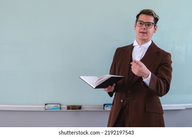 A School Teacher In Front Of The Blackboard. A Teacher Giving A Class With A Book In Hand. Teacher Explaining A Topic To His Students. High Quality Photo