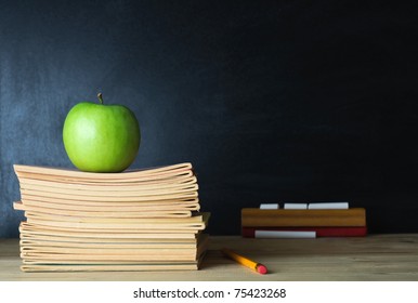 A school teacher's desk with stack of exercise books and apple in left frame. A blank blackboard in soft focus background provides copy space. - Powered by Shutterstock