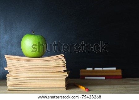 A school teacher's desk with stack of exercise books and apple in left frame. A blank blackboard in soft focus background provides copy space.