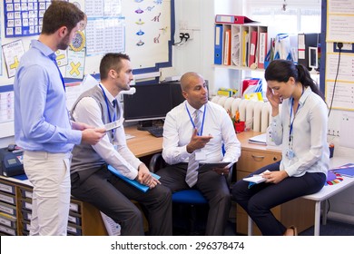 School Teachers Gather In A Small School Office For A Chat. They Look Serious. A Woman And Three Men Group Together. The Woman Holds Her Hand To Her Head.