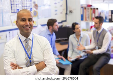 School Teachers Gather In A Small School Office For A Chat. They Look Happy. The Focus Is On The Man Who Stands In The Foreground With The Others Sitting Out Of Focus Behind. He Smiles At The Camera.