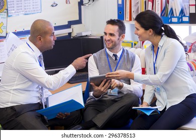 School teachers gather in a small school office for a chat. They look happy. A woman and two men group together. A man holds a digital tablet