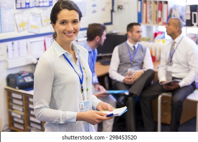 School Teachers Gather In A Small School Office For A Chat. They Look Happy. The Focus Is On The Woman Who Stands In The Foreground With The Men Sitting Out Of Focus Behind. She Smiles At The Camera.