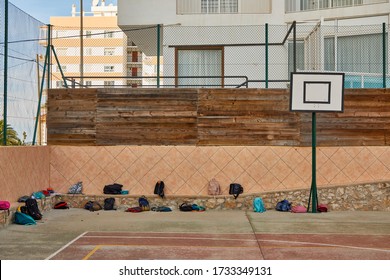 School Yard Full Of Backpacks On The Floor With A Broken Basketball Hoop, With A Wooden Wall And Buildings In The Background