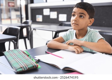 In school, young biracial boy sitting at a desk in a classroom, looking thoughtful. He has dark hair, wearing a light blue t-shirt, with a notebook and pencils in front, unaltered. - Powered by Shutterstock