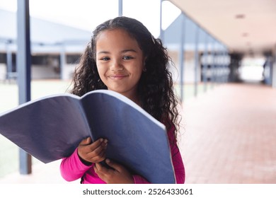 In school, young biracial female student holding a book, standing outdoors. She has curly brown hair, dark eyes, and is wearing a pink top, unaltered. - Powered by Shutterstock