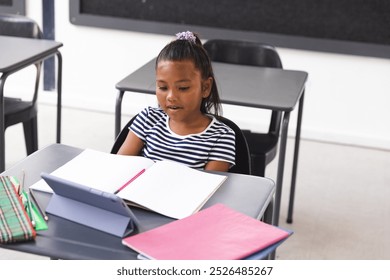 In school, young biracial female student reading a book at her desk in a classroom. She has brown hair tied up, wearing a striped shirt, and a tablet on desk, unaltered. - Powered by Shutterstock