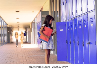 In school, young biracial female student holding books, standing by lockers outdoors with copy space. She has long, dark hair, wearing a floral dress and carrying an orange binder, unaltered. - Powered by Shutterstock