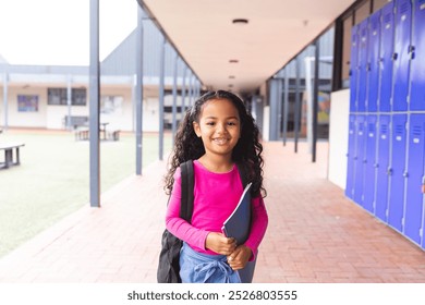 In school, young biracial female student holding a tablet stands smiling. She has curly dark hair, light brown skin, and is wearing a pink top, unaltered - Powered by Shutterstock