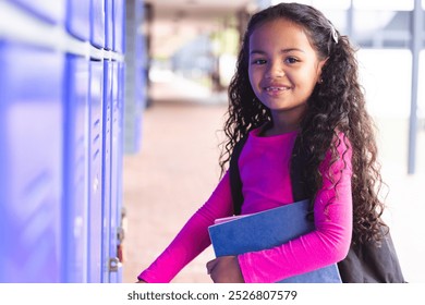 In school, young biracial female student holding books, standing by lockers outdoors. She has long curly dark hair, wearing a bright pink top and jeans, unaltered. - Powered by Shutterstock