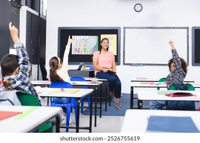 In school, young biracial female teacher smiles at students raising hands in class. She has black hair, wearing a pink top, and holding a recycle poster, learning, education, unaltered - Powered by Shutterstock