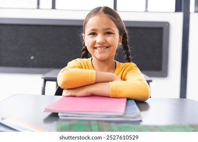 In school, young biracial girl with braided hair sitting at desk in classroom, smiling. She wearing a yellow top, with notebooks on desk, looking at camera, unaltered - Powered by Shutterstock