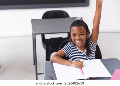 In school, young biracial girl sitting at a desk in a classroom raising hand with copy space. She has dark hair tied up, wearing a striped shirt, and holding a pink pen, unaltered. - Powered by Shutterstock