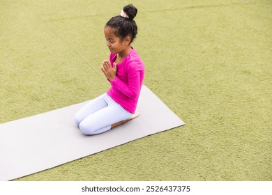 In school, young biracial girl wearing pink is practicing yoga outdoors. She has her hands together, eyes closed, and is sitting on a mat, unaltered. - Powered by Shutterstock