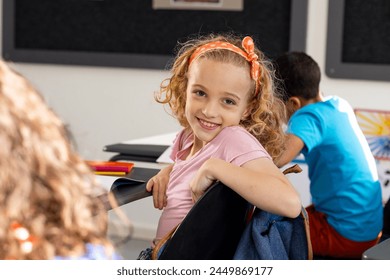 In school, young Caucasian girl with curly hair is smiling, sitting at a desk in a classroom. She has light brown hair with an orange hairband and wears a pink shirt, unaltered. - Powered by Shutterstock