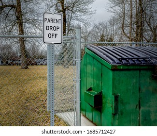 School Zone Parent Drop Off Sign Next To A Rubbish Dumpster In Front Of A Fence And  Trees And Houses In The Background