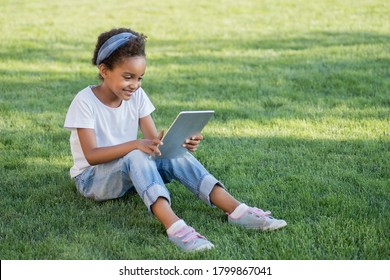 School-age Child With An Electronic Device.  Outside. Mixed Race Girl Sits On A Green Lawn With Tablet. Emotional Face. Concept Of Online Communication, Learning And Playing With The Help Of Gadgets. 