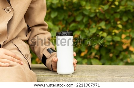 Schoolboy (child) sits on bench and drinks hot tea from thermos outside in park, wears warm beige coats (jackets) Autumn beverage, fall leaves season. Horizontal. Green yellow background. Copy space