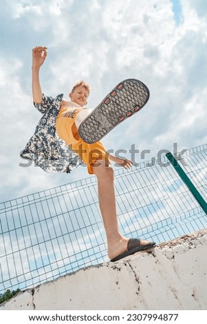 Schoolboy with delighted and smiling expression on high pier against blue sky with clouds. Boy raises foot upward enjoying spending summer holidays at seaside