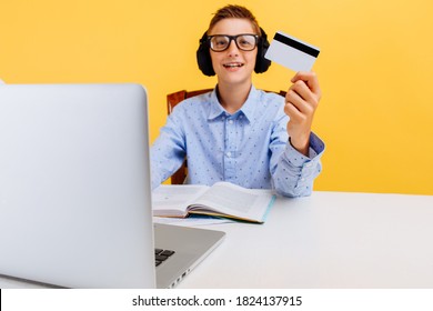 Schoolboy Guy Is Sitting At A Table With A Laptop And Holding A Credit Card On A Yellow Background. Online Shopping, Coronavirus