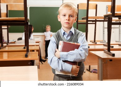 Schoolboy Is In The Half Empty Classroom After Classes. He Holds Copybooks In His Hands. He Is Bored. Teacher Is Teaching Schoolgirl Behind Him.